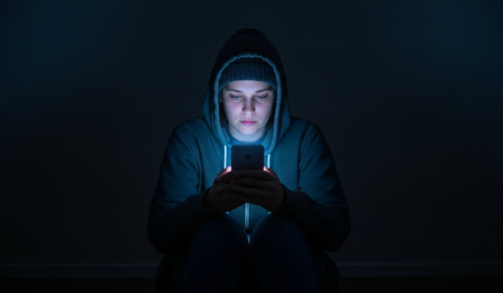 A lone person sitting in a dark room, face lit only by the blue glow of a smartphone screen